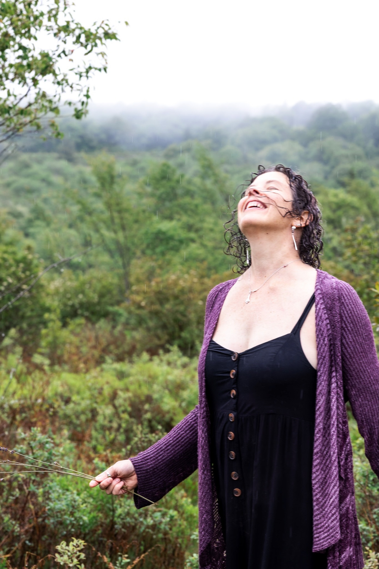 Rachelle laughing skyward in the rain, wearing a purple cardigan, with misty hills behind her.